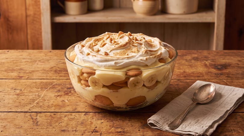 Banana pudding in a glass bowl with meringue and cookie crumbles on a wooden table.
