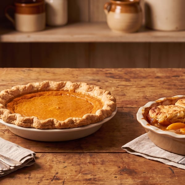 A pumpkin pie and a peach cobbler on a rustic wooden table with pottery in the background.